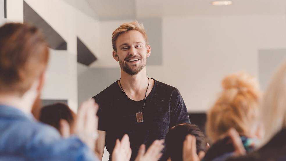 Ein Mann in einem schwarzen Hemd steht in einem Raum voller Menschen, die klatschen.