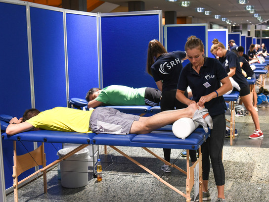 Foto: Im Vordergrund liegt ein Marathon-Läufer auf einer Liege und wird von einer Physiotherapeutin massiert.