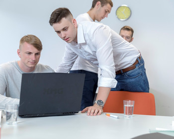 Zwei Teilnehmer sitzen vor einem PC. Im Hintergrund befinden sich ebenfalls zwei junge Männer.