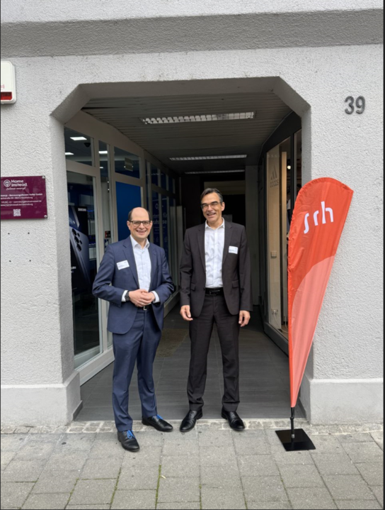 Zwei Männer stehen vor einem Gebäude, neben einem Schild mit der Aufschrift "srh".
