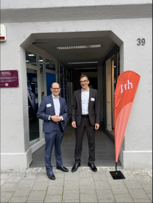 Zwei Männer stehen vor einem Gebäude, neben einem Schild mit der Aufschrift "srh".