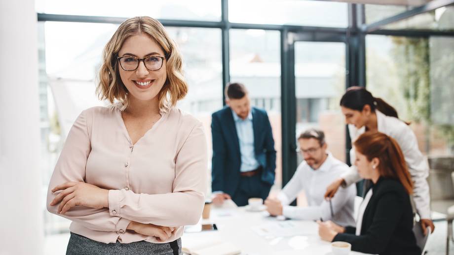 Eine lächelnde Frau mit Brille steht selbstbewusst vor einem Meeting-Tisch mit mehreren besprechenden Personen.