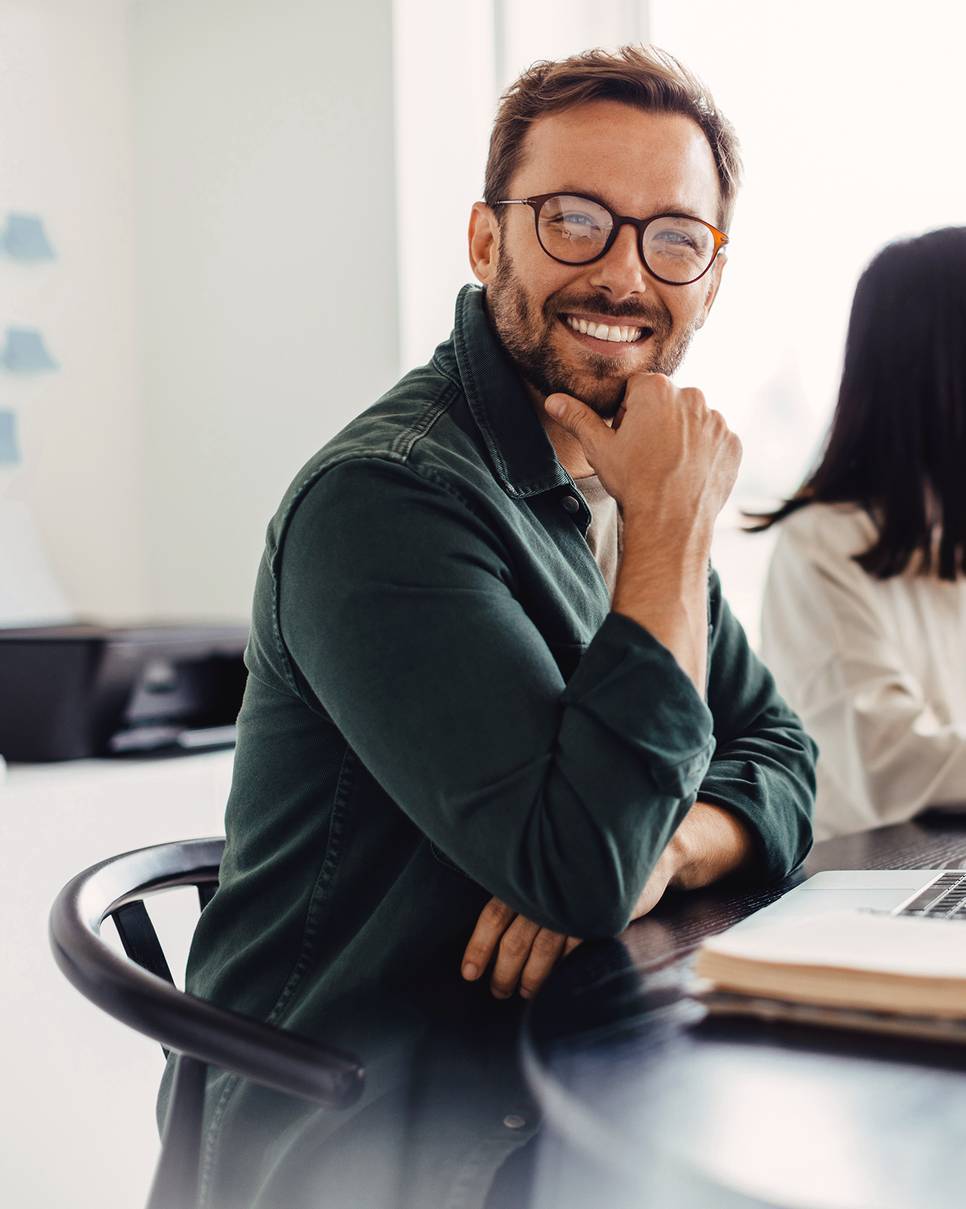 Ein junger Mann sitzt an einem PC-Arbeitsplatz und schaut lächelnd zu anderen Personen