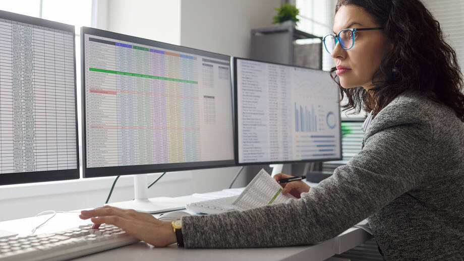 Eine Frau mit Brille und Blazer sitzt an einem Arbeitsplatz mit drei Monitoren an denen Statistiken und Tabellen zu sehen sind.