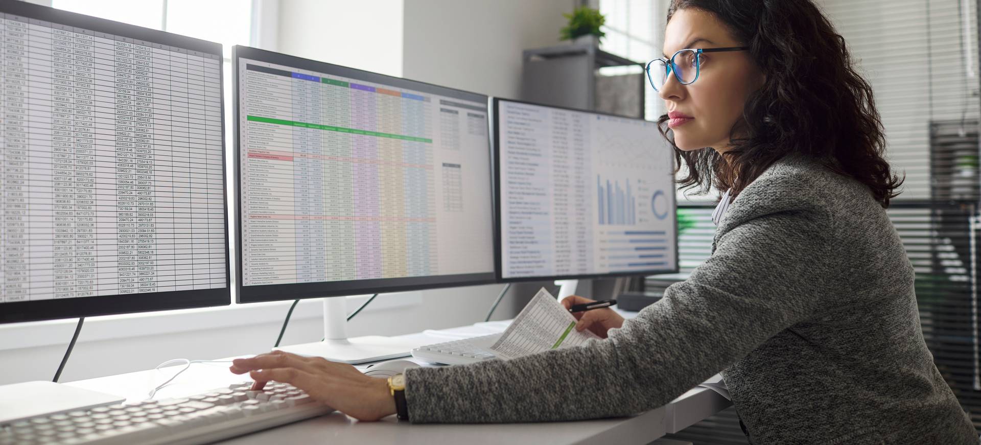 Eine Frau mit Brille und Blazer sitzt an einem Arbeitsplatz mit drei Monitoren an denen Statistiken und Tabellen zu sehen sind.