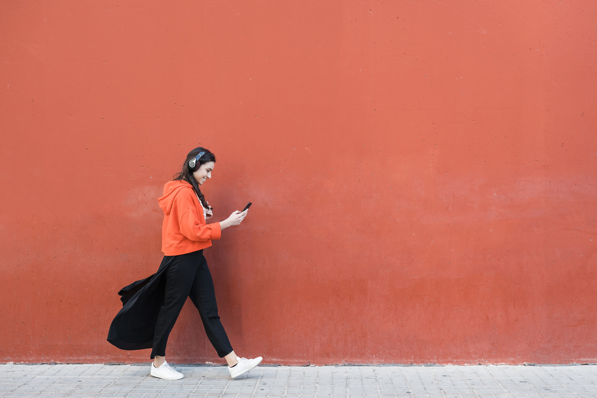 Frau läuft an roter Wand draußen vorbei während sie auf das Handy schaut und Kopfhörer trägt