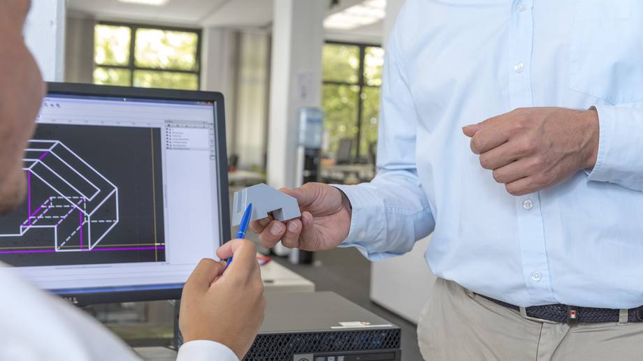 Zwei Menschen sitzen vor einem Bildschirm und zeigen auf einen Gegenstand..