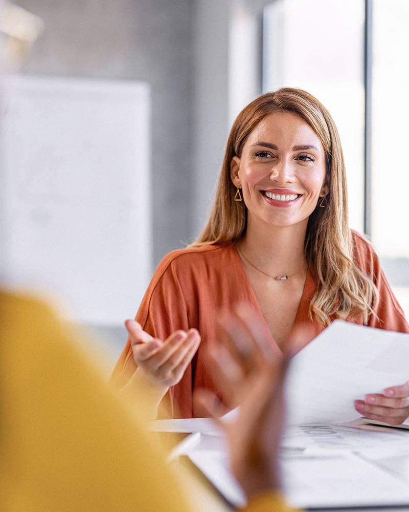 Zwei Frauen sitzen sich an einem Arbeitsplatz gegenüber, die dem Fotografen zugewandte Person lächelt und hält Dokumente in der Hand.