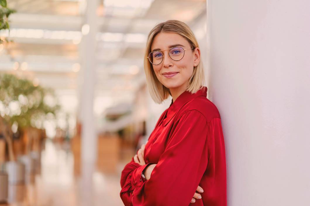 Frau ist mit Rücken an Türrahmen angelehnt und lächelt in die Kamera. Die Sicht ins Zimmer dahinter ist verschwommen