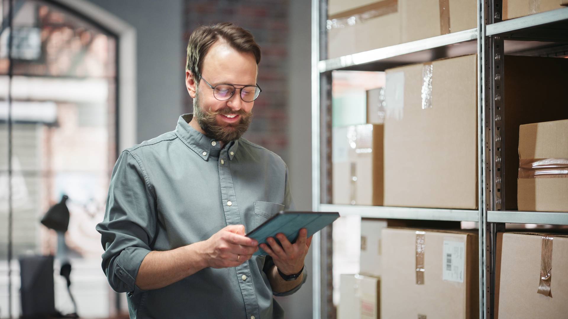 Ein Mann mit Brille und legerem Hemd steht vor einem Regal mit Paketen und schaut in sein Tablet.