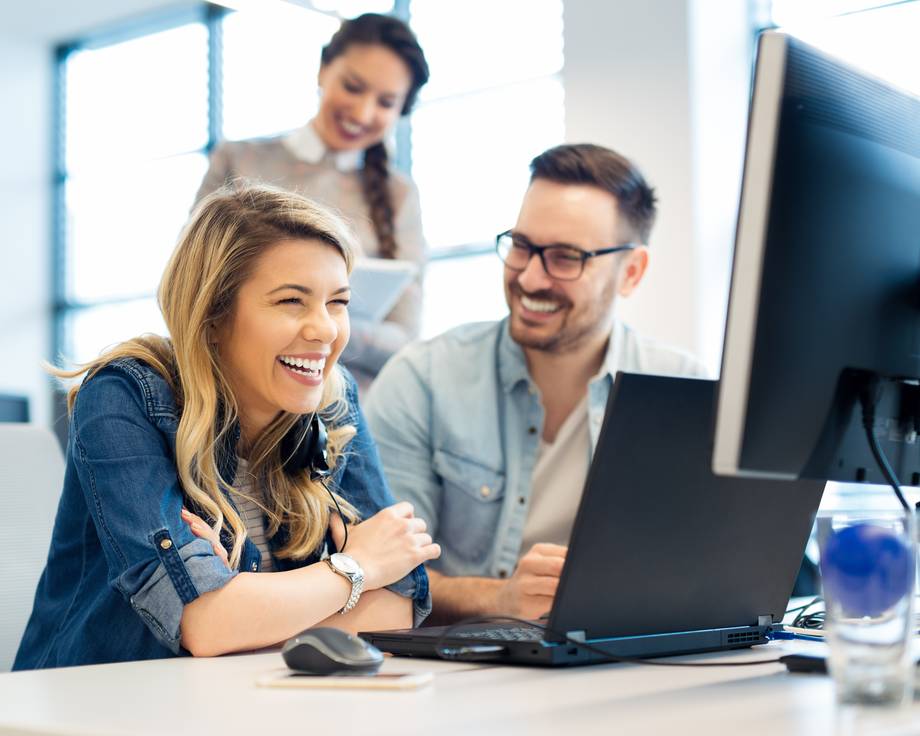 Drei Personen arbeiten an einem Schreibtisch mit Computern, eine Frau lacht