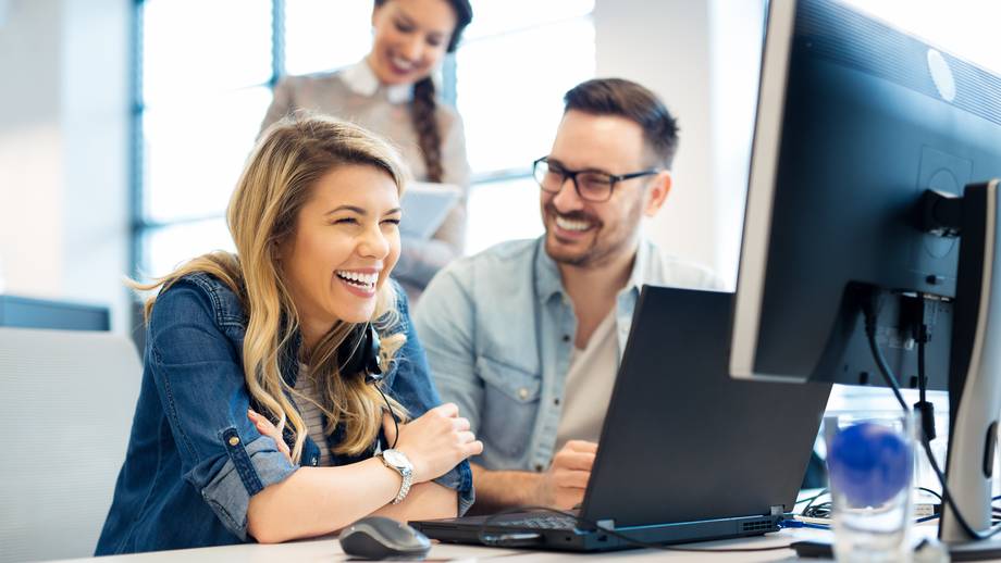 Drei Personen arbeiten an einem Schreibtisch mit Computern, eine Frau lacht