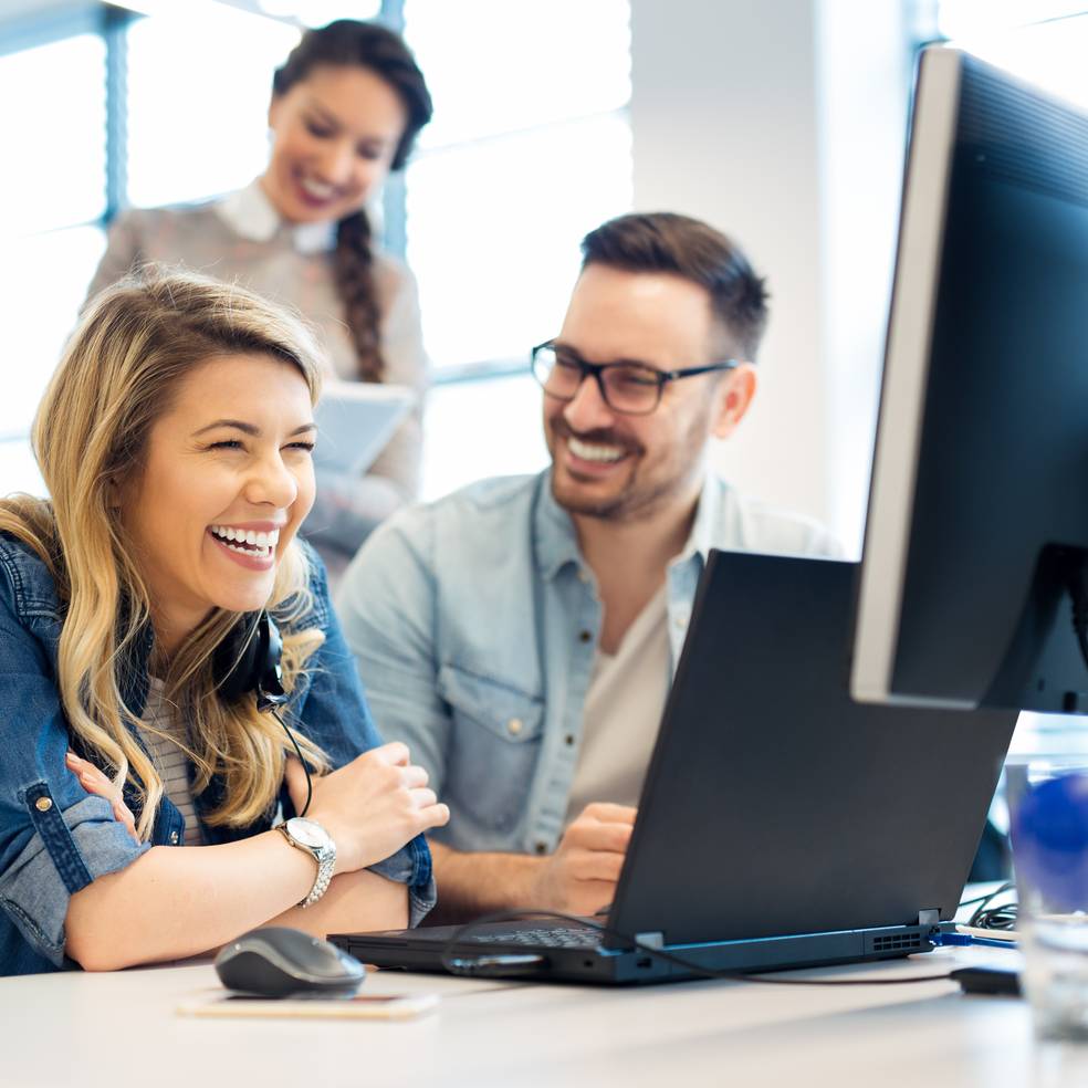 Drei Personen arbeiten an einem Schreibtisch mit Computern, eine Frau lacht