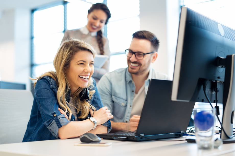 Drei Personen arbeiten an einem Schreibtisch mit Computern, eine Frau lacht