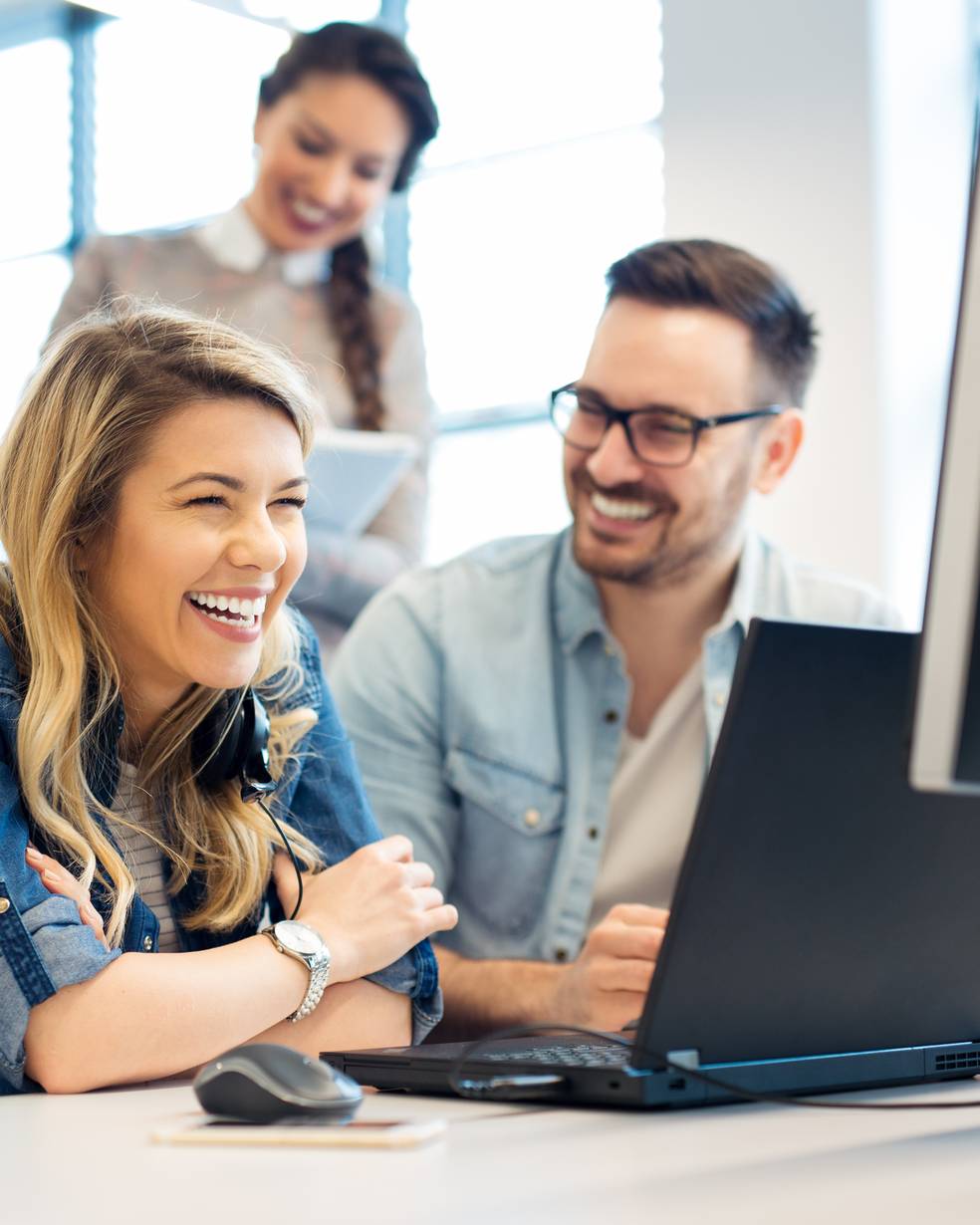 Drei Personen arbeiten an einem Schreibtisch mit Computern, eine Frau lacht