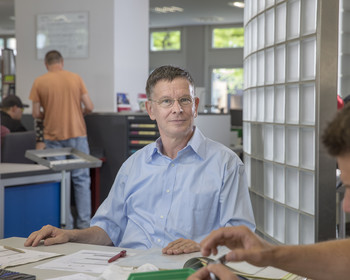 Teilnehmender sitzt am Tischt lächelt, im Hintergrund und rechts arbeiten anderer Teilnehmer an Aufgaben