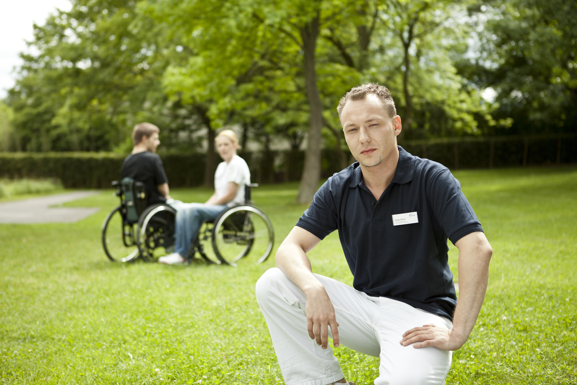Pfleger kniet auf Wiese, im Hintergrund unterhalten sich zwei Rollstuhlfahrer